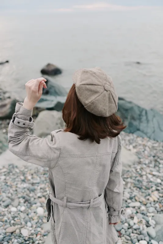Woman in beige coat and matching hat stands on rocky beach near calm sea, looking at water. Autumn outdoor scene with peaceful mood and natural coastal landscape background.