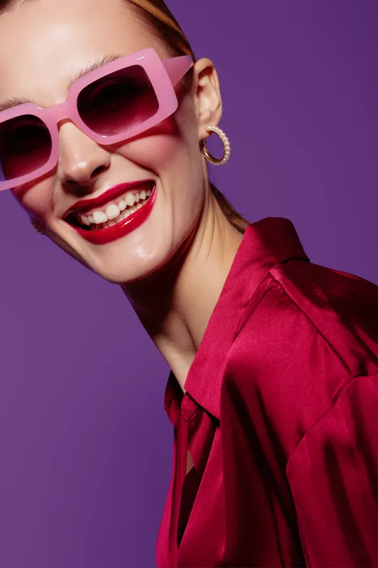 Smiling woman with pink sunglasses and red lipstick wears red satin blouse and gold hoop earrings on purple background. Concept of fashion, beauty, and joyful lifestyle.