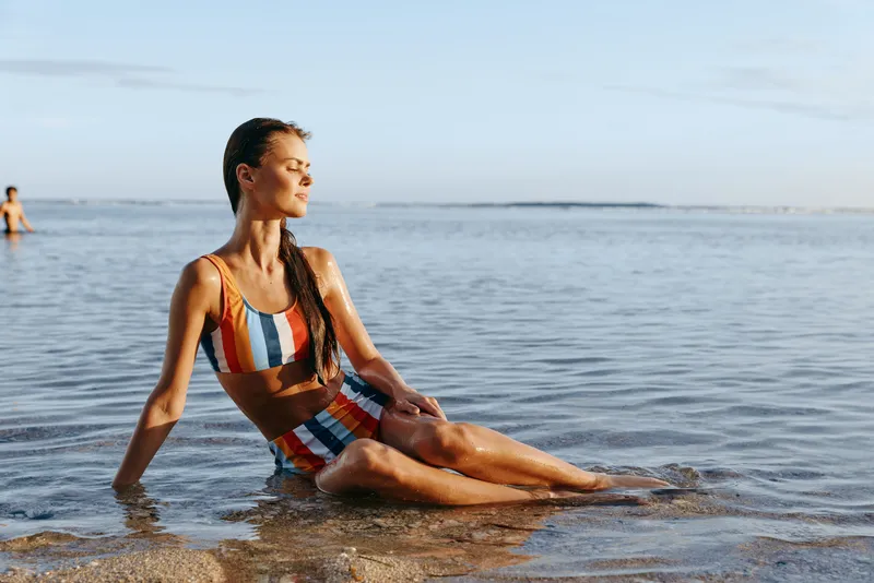 Woman on beach ocean yoga relaxation summer mindfulness serene posture, sitting in shallow sea water, waves and horizon in background, peaceful morning sunlight