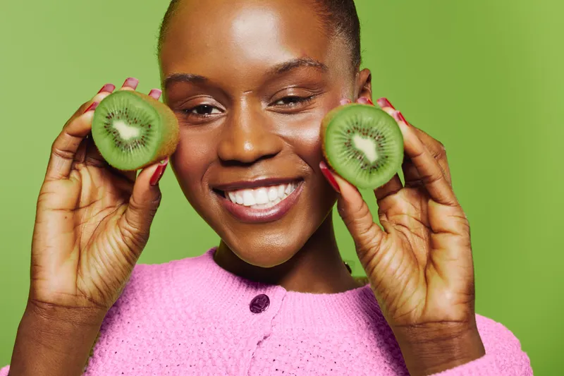 Smiling young black woman with dark skin holds two halves of fresh kiwi fruit near face against green background. Concept of healthy lifestyle, fresh fruit, and natural beauty.