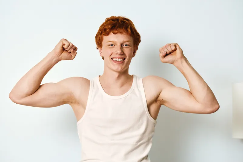 Young man with red hair and white tank top flexing muscles with happy smile against light background in casual style and confident pose.
