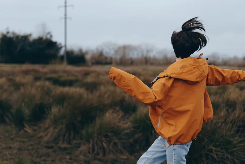 Child outdoor lifestyle in a grassy field, wearing an orange jacket with hood, arms open wide, cloudy evening sky, nature scenery, child happiness, back view, rural calm park walk