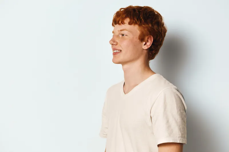 Young man with red hair and pale skin standing in white t shirt against plain light background looking to side with slight smile.