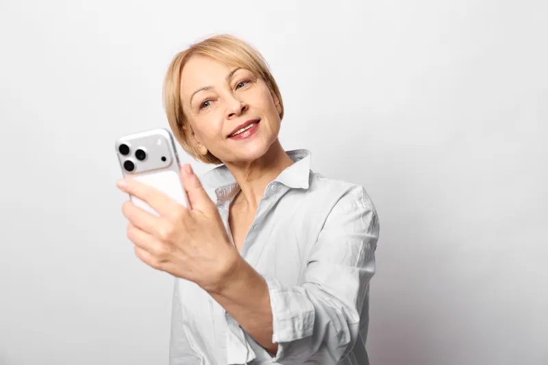 Woman in studio portrait holding smartphone taking selfie, mature confident face, video call, light background, communication technology, relaxed media pose