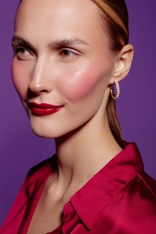 Close-up of a woman with glossy red lips and pink blush wearing a red blouse against a purple background. Concept of beauty, makeup, fashion, and skincare.