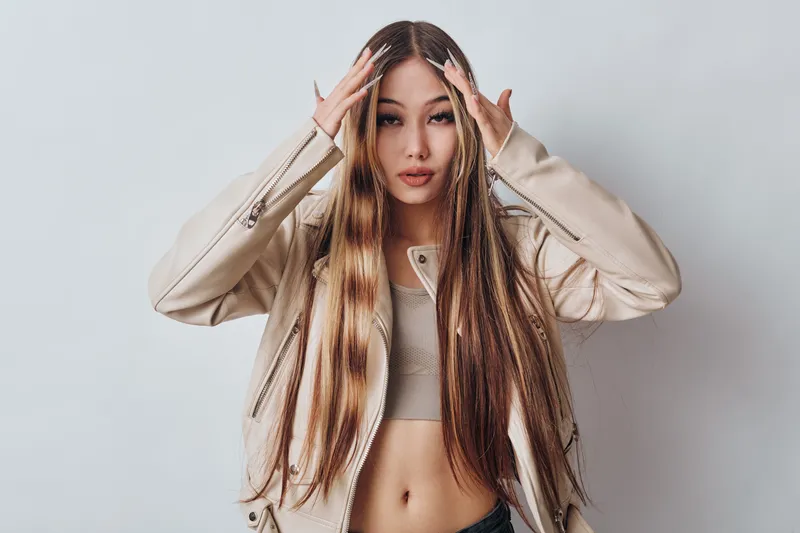 Young woman with long hair wearing beige jacket and crop top posing with hands on head against white background in studio portrait fashion style.