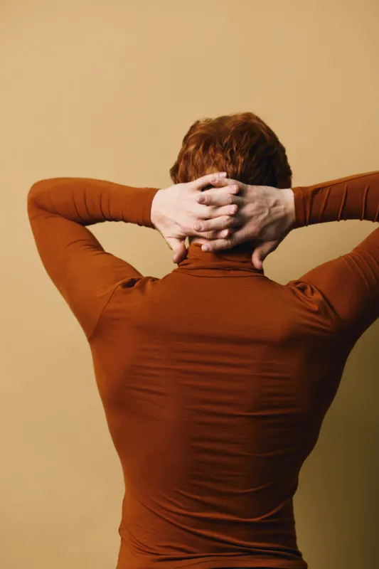 Person with red hair wearing brown turtleneck stretching arms behind head against beige background. Relaxation, casual style, and calm mood with focus on upper back and posture in studio.