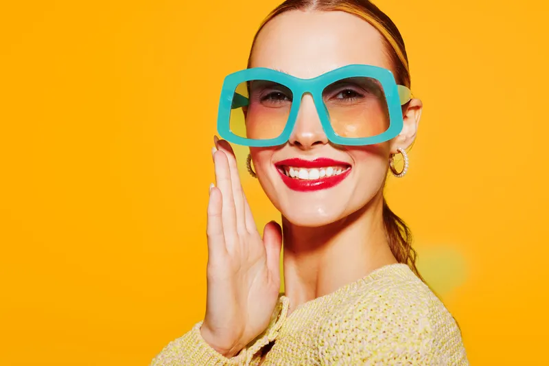 Smiling woman with large blue glasses and red lipstick poses on bright orange background. She wears yellow sweater and gold hoop earrings. Concept of fashion, style, and vibrant portrait.