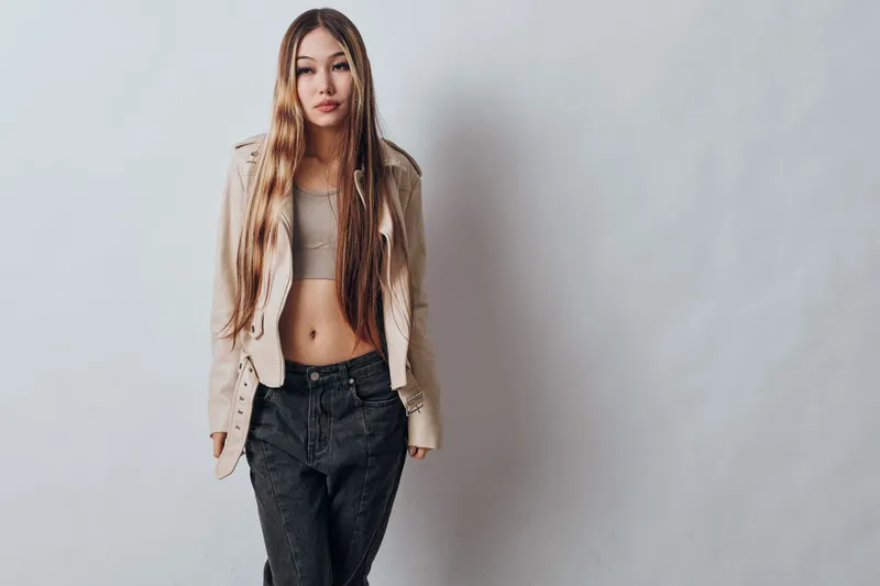 Young woman with long braided hair wearing casual crop top and beige jacket posing confidently with hands in pockets against gray studio background.