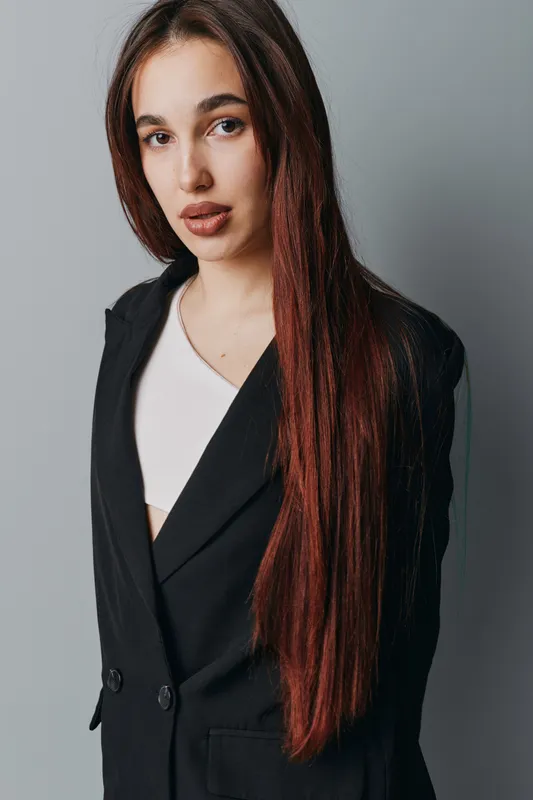 Young woman with long straight hair wearing black blazer and white top standing against neutral background for portrait and fashion concept. Female model with natural makeup and confident expression.