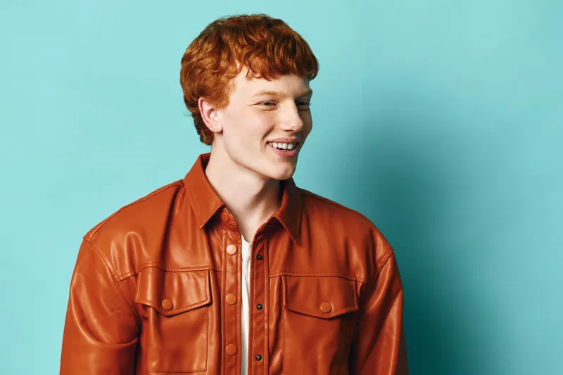 Young man with red hair wearing orange leather jacket smiling and looking sideways against plain blue background. Casual style, happy male portrait with natural expression and fashion look.