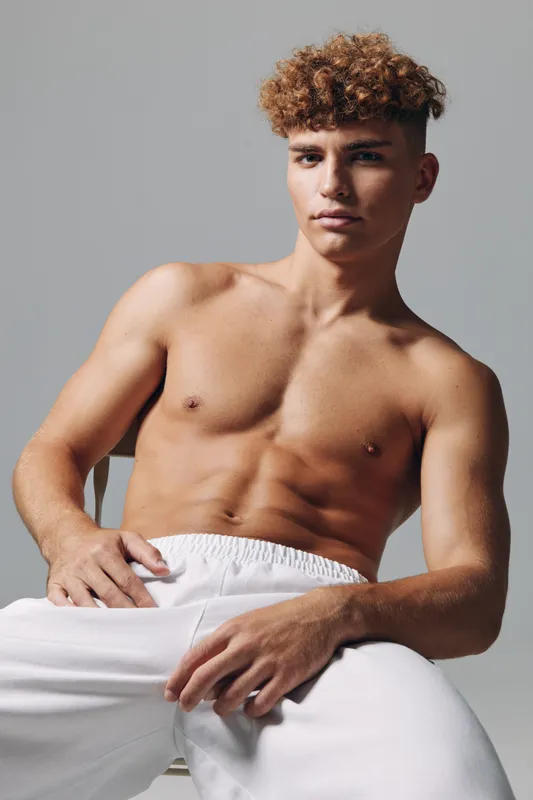 Young man with curly hair and muscular torso posing shirtless in white pants against gray background. Fitness, confidence and casual style with natural light and relaxed posture.