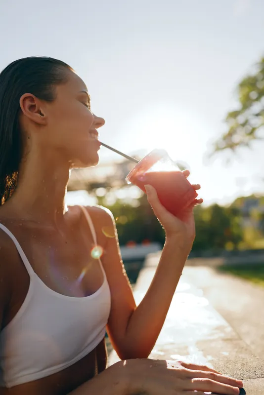 Woman in a white top drinks juice outdoors on a sunny day, capturing a casual lifestyle moment focused on hydration and wellbeing under warm natural light
