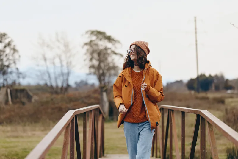 Woman lifestyle outdoors bridge, smiling woman in orange jacket and knit beanie, walking on wooden bridge in autumn fields, travel mood, countryside calm, relaxed, natural daylight
