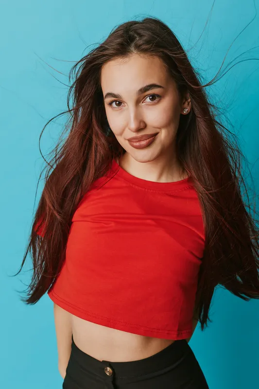 Young woman with long hair wearing red crop top and black pants posing confidently against blue background. Portrait of smiling female with natural makeup and casual style.