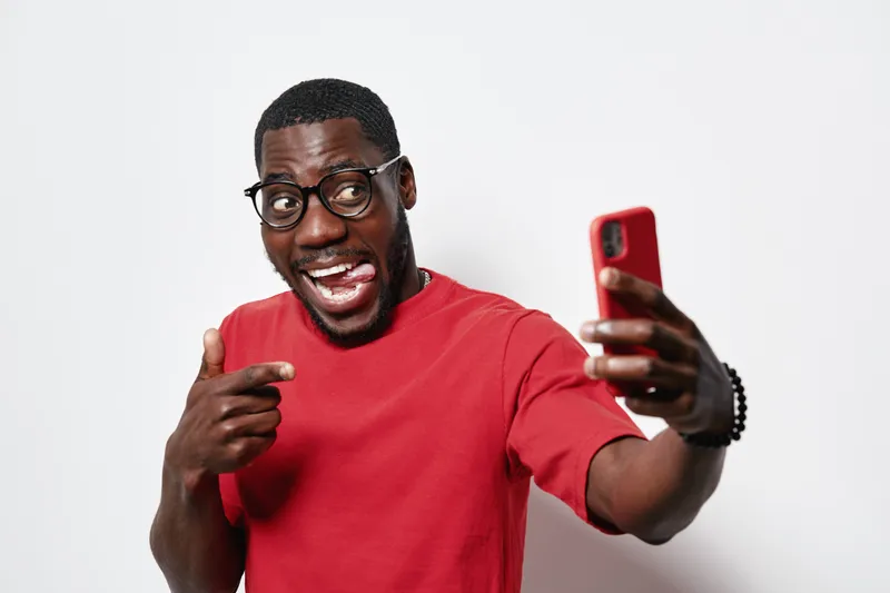 Black man with dark skin and glasses wearing red shirt taking selfie with smartphone, smiling and pointing finger, isolated on white background with happy expression.