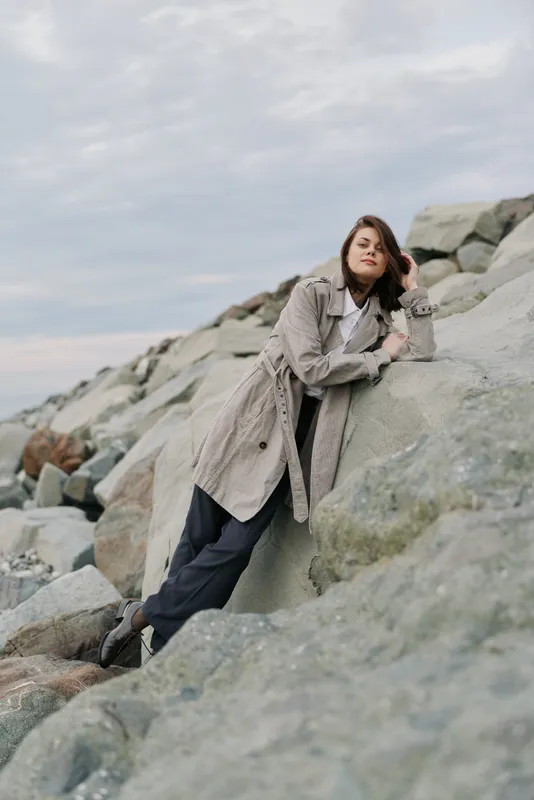 Young woman in long coat leans on large rocks by cloudy sky. Casual outdoor fashion photo with natural light and relaxed pose on rocky landscape near sea.