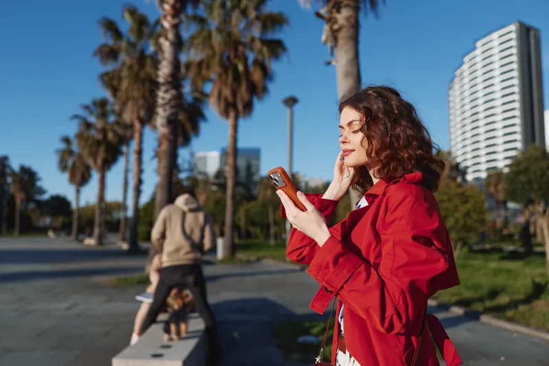 Young woman outdoors using smartphone, casual red jacket, curly hair, sunny day, urban park setting, relaxed expression, modern city background, palm trees.