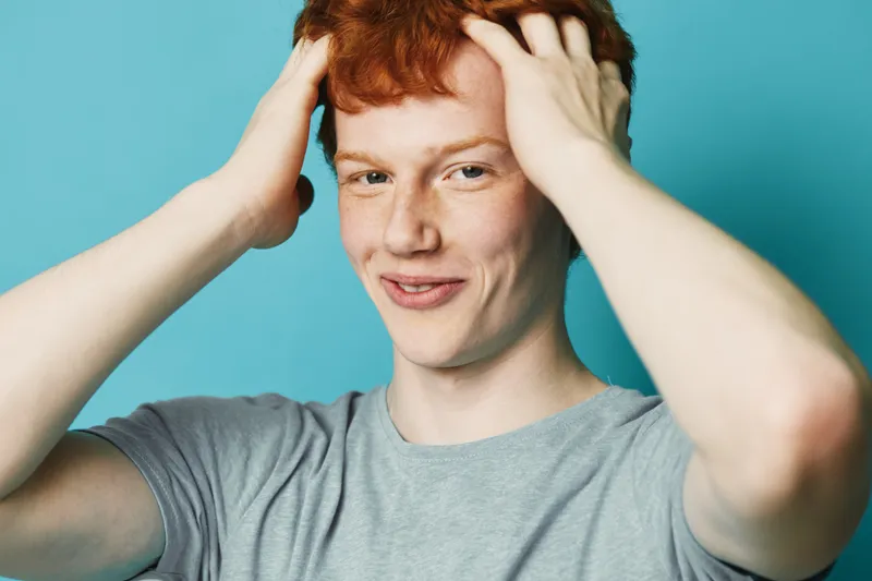 Young man with red hair and freckles wearing gray t shirt holding head with hands against blue background. Portrait of confident smiling male with casual style and natural look.