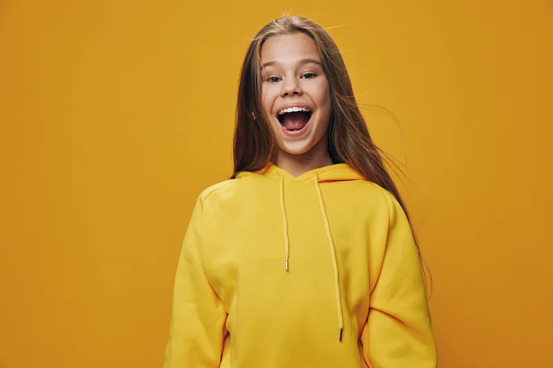 Girl in yellow hoodie smiling with open mouth on bright orange background for happiness, emotion, youth and casual fashion portrait of cheerful child with long hair and positive vibe.