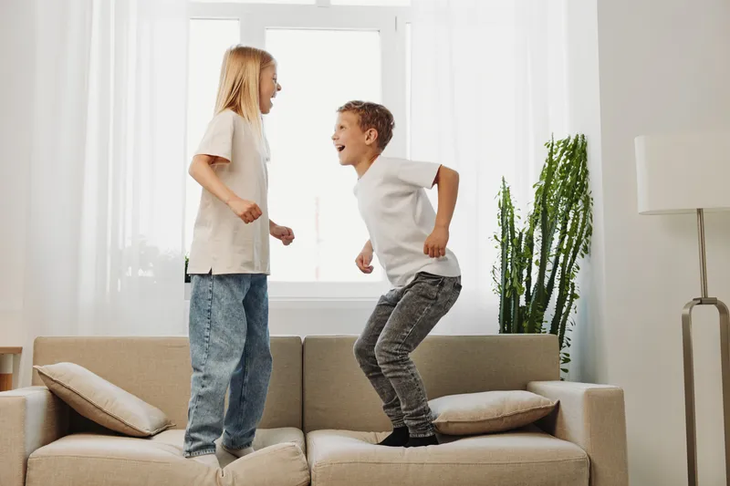 Two children jumping on beige sofa in bright living room with white curtains and green plants, enjoying playful moment together indoors.