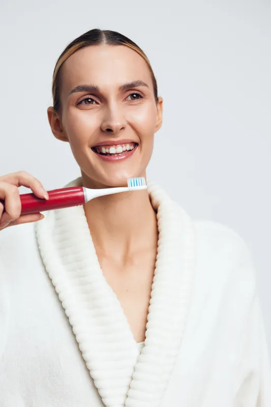 Smiling woman in white robe holding red toothbrush with toothpaste, bright studio lighting, clean background, dental hygiene concept, healthy teeth and fresh breath