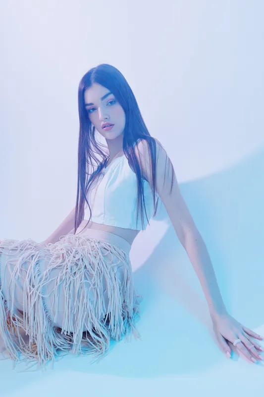 Young woman with long hair in a white top and fringed skirt poses sitting on the floor in soft blue studio light. Concept of beauty, fashion, and elegance with modern style.