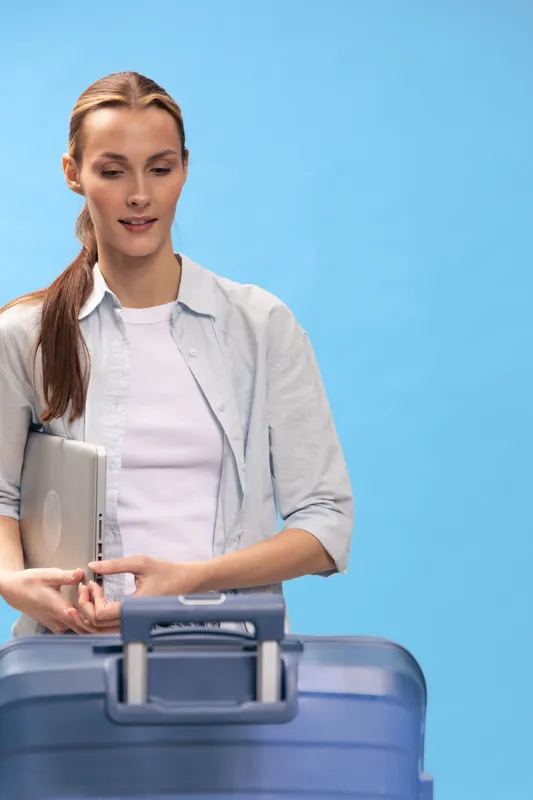 Young woman with laptop stands behind blue suitcase on blue background. Travel concept with casual outfit and confident expression preparing for journey or business trip.
