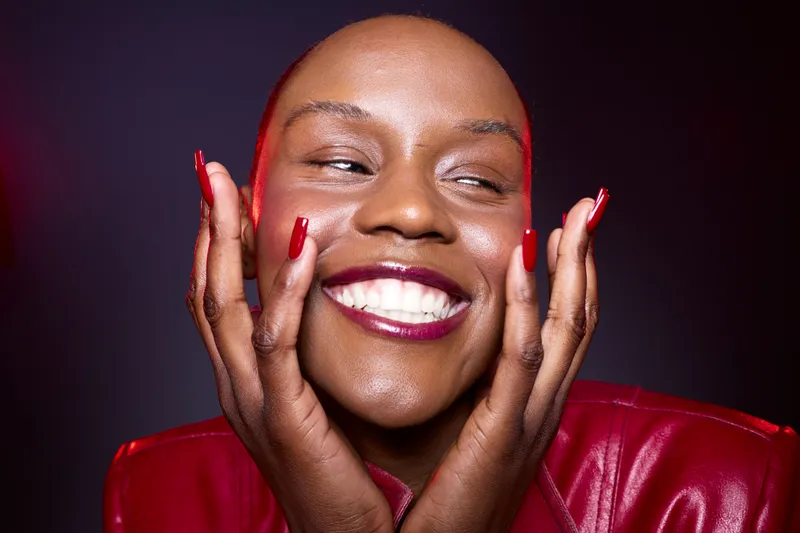 Close-up of happy African woman with dark skin smiling brightly, hands on face. She wears red lipstick and red jacket, studio portrait with dark background. Concept of joy and beauty.