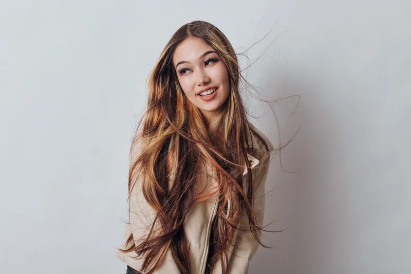 Young woman with long hair posing and smiling against studio background looking to the side wearing casual clothing with natural makeup and joyful expression