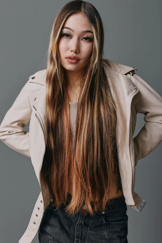 Young woman with long straight hair wearing jacket and black pants standing with hands on hips on gray background. Casual fashion look and confident pose in studio lighting.