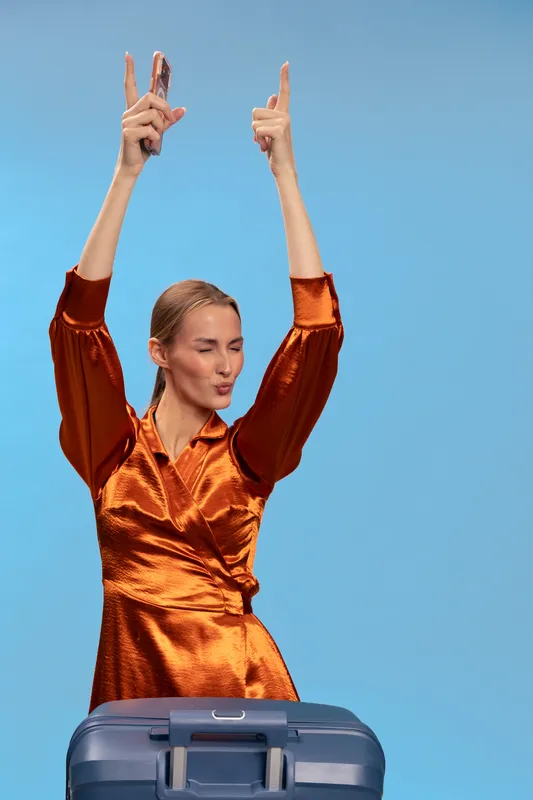 Excited woman in orange satin dress holds smartphone and raises hands against blue sky. Travel concept with suitcase, happiness, freedom, and celebration in bright daylight.