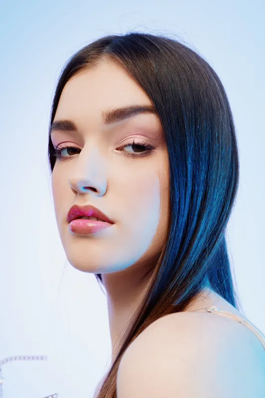 Close-up portrait of a young woman with long dark hair and pink makeup looking over her shoulder. Soft lighting highlights her smooth skin and beauty. Concept of fashion and elegance.