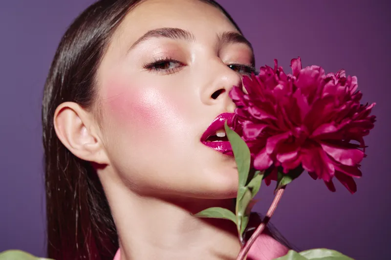 Young woman with glossy fuchsia lipstick and pink shimmer makeup holds deep red flower near her face on purple background. Creative beauty and fashion portrait concept.