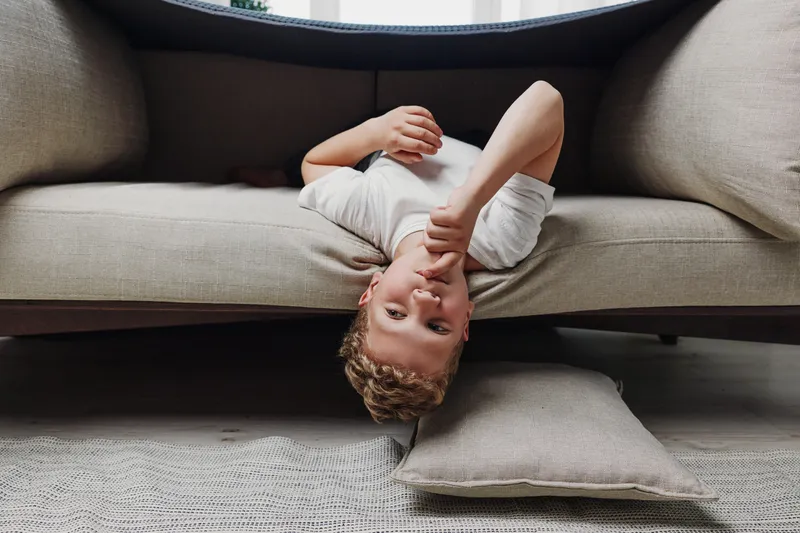 A young boy lies upside down on a beige sofa with a cushion on the floor, looking thoughtfully at the camera in a cozy living room. Concept of childhood, play, and relaxation.