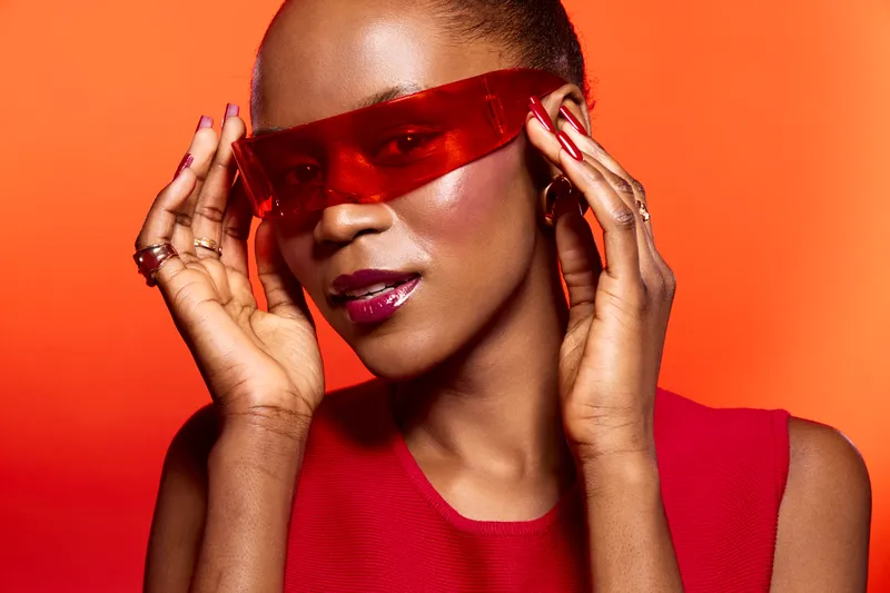 Stylish black woman with dark skin wears red futuristic glasses and red sleeveless top. Close-up portrait on orange background. Concept of fashion, beauty, and modern accessories.