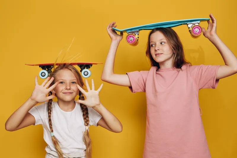 Two smiling girls holding skateboards above their heads against yellow background. They wear casual clothes and show playful expressions. Concept for childhood fun.