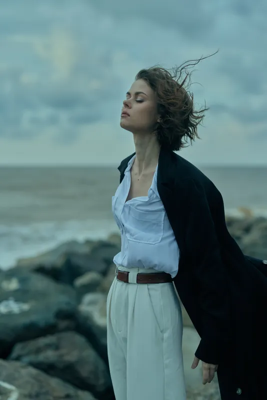 Windy seaside outdoor fashion scene with ocean breeze and moody atmosphere, a wind swept figure in a longcoat and white shirt stands among rocks by the shore, calm and contemplative