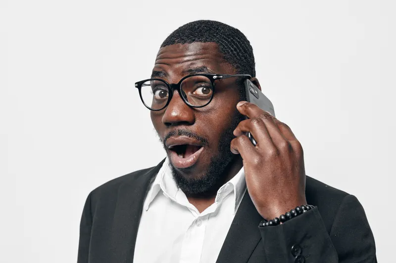 African American man with dark skin wearing glasses and black suit talking on smartphone with surprised expression on white background. Communication, technology and business concept.