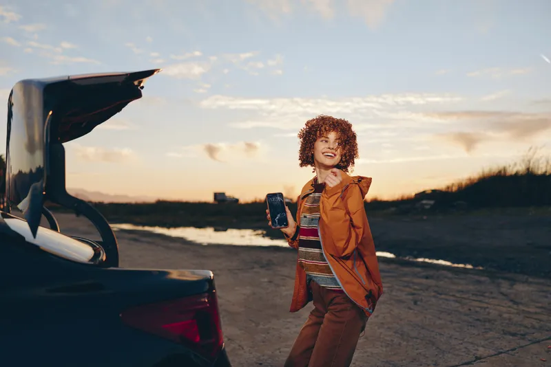 Woman lifestyle portrait outdoors at sunset, standing on a road by a parked car with an open trunk, curly haired woman holds a bag, travel mood, warm golden light