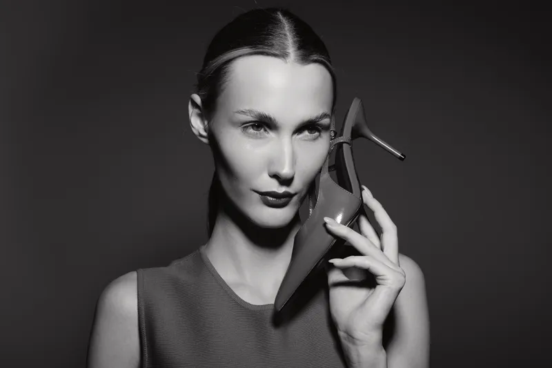 White woman with fair skin and slicked hair holding high heel shoe near face in black and white portrait studio photo.