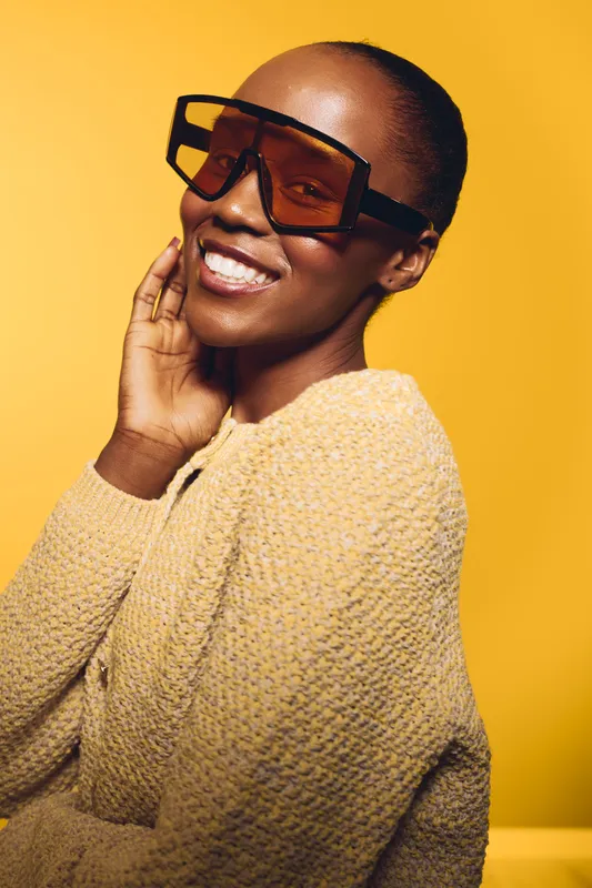 Smiling dark skinned African woman wears large sunglasses and beige knitted sweater, posing on yellow background. Concept of fashion, beauty, confidence, and modern lifestyle portrait.