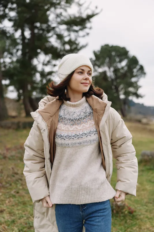 Woman wearing a warm coat and knit sweater stands outdoors in a crisp countryside setting exuding cozy style and confidence as trees blur softly in the background