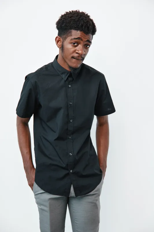 Young black man with dark skin wearing black shirt and gray pants standing with hands in pockets on white background. Casual style, confident pose and thoughtful expression.