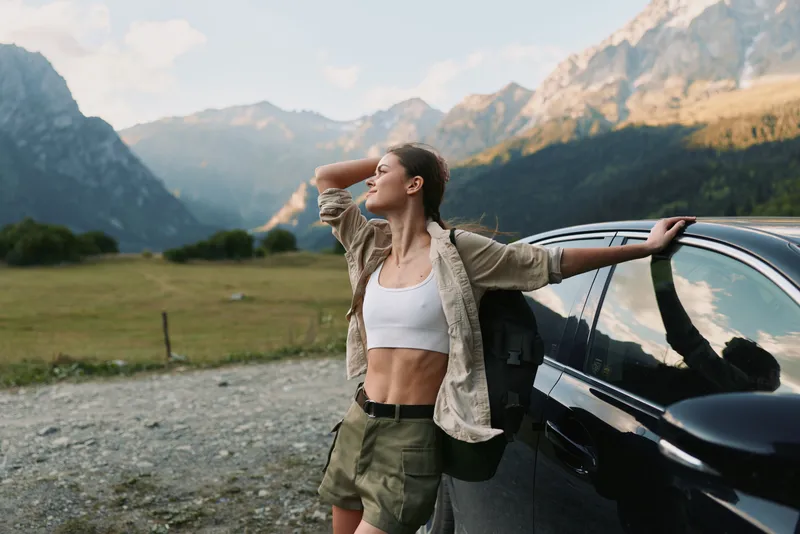 A woman stands beside a dark car in a rocky landscape, wearing a white crop top and beige jacket, arms raised, enjoying sunshine and freedom in the mountains, conveying outdoor lifestyle.