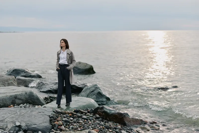 Woman stands on rocky shore near calm sea under cloudy sky. She wears casual clothes and looks thoughtfully into the distance. Concept of solitude, nature, and contemplation.