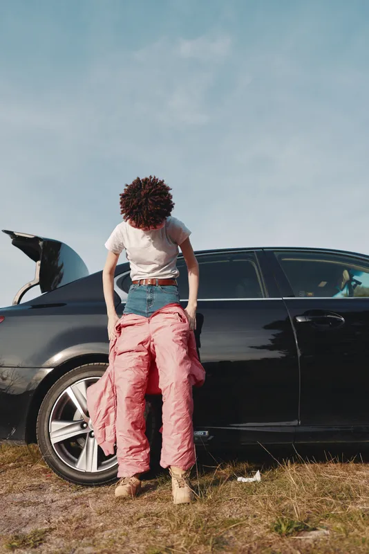 Person beside a car with the hood open performing roadside inspection and repair during daytime, casual clothing, dusty ground, open landscape, wheel and tools visible, highlighting vehicle