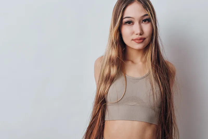 Young woman with long straight hair and neutral expression wearing beige crop top standing against light background for beauty, fashion and lifestyle portrait.
