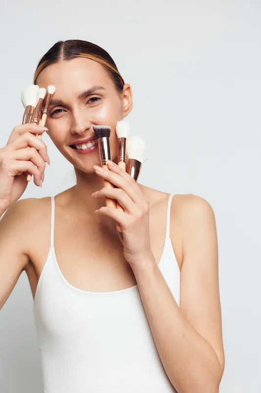 A smiling young woman in a white tank top holds multiple makeup brushes near her face against a plain background. Concept for beauty, skincare, and makeup tools.