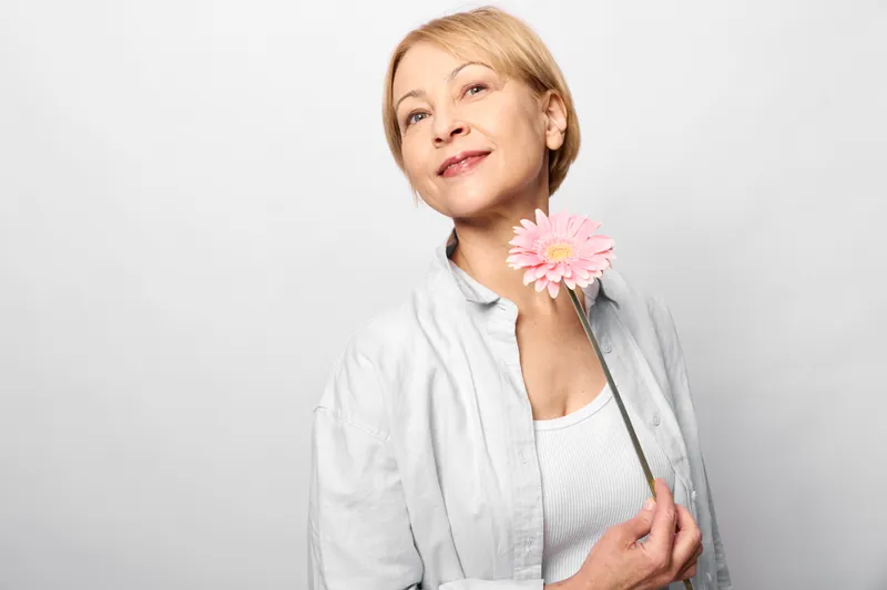 Portrait of an elderly woman holding a pink flower against a white studio background, with calm smile and tenderness, soft expression and light suggest serenity and care